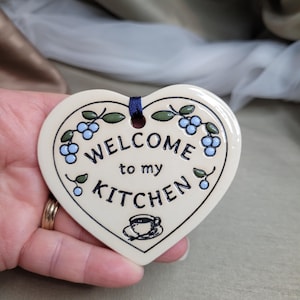May include: Cream-coloured heart-shaped kitchen sign with the words "WELCOME to my KITCHEN" in black. It features blue berries and green leaves around the edge. A blue ribbon is attached for hanging. The sign is made of ceramic.
