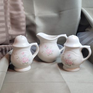 Può includere: Una crema in ceramica bianca e due saliere con un motivo floreale a rose. Ogni pezzo ha un manico ed è decorato con rose rosa e foglie verdi. Il set è per la tavola da pranzo.
