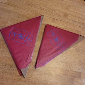 Op de afbeelding: Twee driehoekige, rode bakzakken met blauwe illustraties en tekst. De zakken zijn gedeeltelijk open en onthullen een kartonnen basis met een liniaalrand. De zakken liggen op een houten oppervlak.