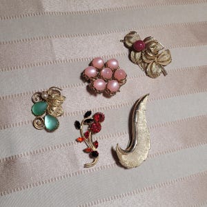 Puede incluir: Una colección de broches vintage de varios diseños. Incluye un broche de filigrana dorado con una piedra rosa, un grupo de perlas de imitación rosas y un broche dorado en forma de hoja. Otros broches presentan detalles en verde y rojo.