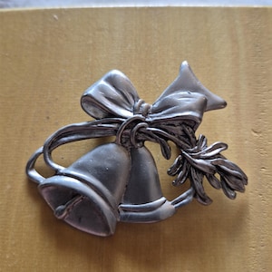 Könnte beinhalten: Silberfarbene Metallbrosche mit zwei Glocken, die mit einer Schleife und einem Zweig aus Blättern verbunden sind. Die Brosche hat ein detailliertes Design und ist wahrscheinlich ein dekoratives Accessoire.