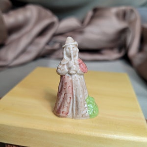 May include: A small ceramic figurine of a person in a long robe and hat. The figurine is mainly beige with red and green details. The figure is standing on a small green base.