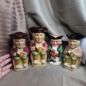 Könnte beinhalten: Vier Keramik-Toby-Krüge in einer Reihe. Jeder Krug zeigt einen Mann mit Dreispitzhut, der einen Bierkrug hält. Die Krüge sind in verschiedenen Farben gehalten. Der Hintergrund ist neutral.