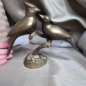 Puede incluir: Escultura de pájaros de color bronce que representa dos aves posadas en una rama. Las aves se miran entre sí, con detalles precisos. La escultura se asienta sobre una base texturizada, dándole un aspecto vintage.