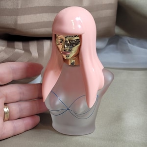 May include: A perfume bottle shaped like a bust with a pink wig and a gold face. The frosted glass bottle has a blue line drawing of a bra. The bottle is on a beige surface.
