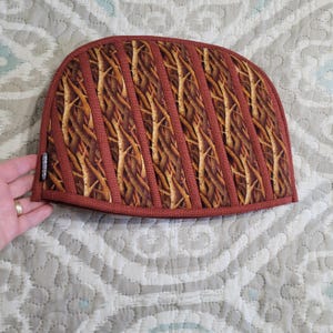 May include: A quilted tea cosy with a curved top and a reddish-brown border. The main fabric features a pattern of brown and tan tree branches. Vertical strips of the reddish-brown fabric create a quilted effect. A small label is visible on the side.