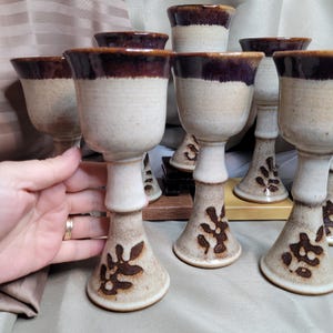 Può includere: Un set di calici in ceramica fatti a mano. I calici hanno un corpo beige con un bordo marrone scuro e un motivo a foglie decorativo. Lo stelo è di colore beige chiaro. I calici sono esposti su una superficie marrone chiaro.