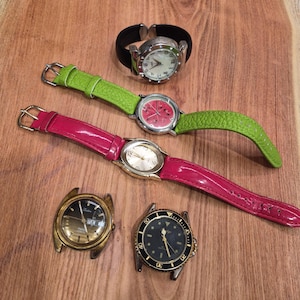 May include: A selection of wristwatches with diverse styles. Features watches with black, green, and pink straps, and watch faces in silver, gold, and black. Some have a classic round design, others have unique shapes.