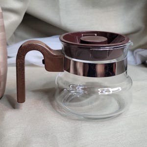 May include: A brown glass coffee pot with a brown plastic lid and handle. The pot has a silver band around the middle.
