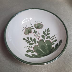 Könnte beinhalten: Weißer Keramiknapf mit grünem Rand und floralem Design. Die Schale hat ein handgemaltes Design aus grünen Blumen und Blättern mit roten Akzenten. Ein dekoratives Stück zum Servieren oder Ausstellen.