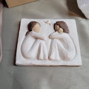 Könnte beinhalten: Eine weiße, geschnitzte Reliefskulptur mit zwei eng beieinander sitzenden Figuren, die ihre Arme um die Knie gelegt haben. Die Figuren haben braunes Haar und tragen weiße Gewänder. Über ihnen ist ein kleiner Stern geschnitzt.