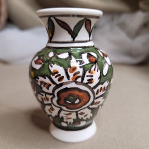 Può includere: Un piccolo vaso in ceramica dipinto a mano con base e bordo bianchi. Il vaso presenta un motivo floreale verde, bianco e marrone. Il corpo del vaso è rotondo, con un collo stretto e una parte superiore svasata. Il vaso è alto circa 10 cm.
