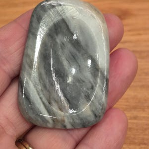 May include: A polished, rectangular stone with rounded edges. The stone has a swirling pattern of grey and white, with a glossy finish. The stone is held in a hand, showcasing its smooth texture and natural colours.