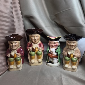 Könnte beinhalten: Vier keramische Toby-Krüge, die jeweils einen sitzenden Mann in traditioneller Kleidung darstellen. Die Krüge haben braune Hüte, und die Figuren tragen Mäntel in Rot-, Grün- und Beigetönen. Sie halten Bierkrüge und lächeln.