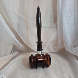 May include: A dark brown glass gavel featuring a long, slender handle and a cylindrical head with ribbed detailing. The handle connects to the head via a silver-coloured metal band, creating a smooth transition between the two distinct glass sections.