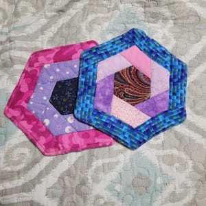 Puede incluir: Dos salvamanteles hexagonales de tela. Uno es rosa con un estampado floral, el otro es azul y morado con un centro de cachemira. Ambos están acolchados con un diseño geométrico.