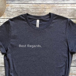 Könnte beinhalten: Ein dunkelgraues T-Shirt mit dem Text "Best Regards," in weiß auf der Vorderseite. Das Shirt liegt flach auf einer Holzoberfläche mit einem Paar schwarzen Sneakers, einem Strohhut, einer Uhr und einer Jeans.