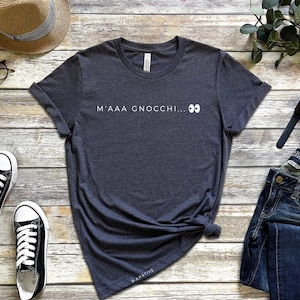 May include: Dark gray t-shirt with the text "M'AAA GNOCCHI... 👀" in white. The shirt is paired with blue jeans, black and white sneakers, and a straw hat. A watch and glasses are also visible.