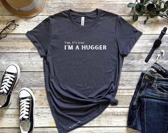 May include: Dark gray t-shirt with the text "Yup, It's true. I'M A HUGGER" in white. The shirt is styled with black and white sneakers, a straw hat, glasses, a watch, and jeans on a wooden surface.