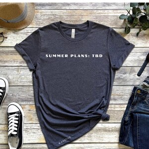 Può includere: Una t-shirt a maniche corte grigio melange scuro con la scritta bianca "SUMMER PLANS: TBD" stampata sul petto. Il capo presenta uno scollo rotondo e una vestibilità standard, presentato come indumento disteso senza altri oggetti inclusi.