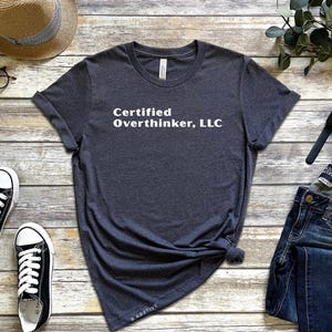May include: A dark grey t-shirt with the text "Certified Overthinker, LLC" in white. The shirt is styled with blue jeans, black and white trainers, a straw hat, and spectacles on a wooden surface.