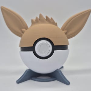 Könnte beinhalten: Eine 3D-gedruckte Pokéball im Evoli-Design. Der obere Teil zeigt Evoli's Kopf mit braunen Ohren und Fell, während der untere Teil ein weißes und schwarzes Ball-Design ist. Er steht auf einem grauen Ständer.