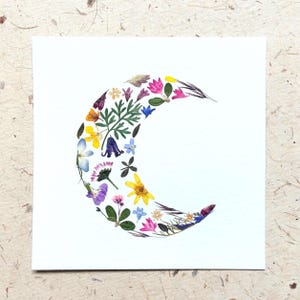Puede incluir: Una tarjeta cuadrada blanca que presenta una forma de luna creciente compuesta por varias flores prensadas y hojas verdes. El arreglo incluye pequeñas flores amarillas, rosas, moradas y azules intercaladas con follaje delicado.