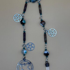 May include: A handcrafted beaded pendant featuring a silver-tone pentagram charm, a castle-shaped charm, and a clear crystal pendant. The beads are a mix of brown, teal, and gray, strung on a silver chain.