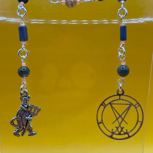 May include: A necklace featuring a silver chain with dark blue and green beads, and a small devil charm holding a trident. The other side has a circular pendant with a symbol inside.