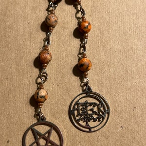 May include: A brown and black beaded necklace with a silver pentagram charm and a silver charm with the word "HEKT" in a stylized font.