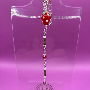 May include: A handmade necklace featuring a red strawberry pendant with white polka dots, green leaves, and a silver chain. The necklace includes red and clear beads, and a silver charm with an intricate design. The necklace is displayed on a clear stand.