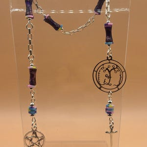 May include: A silver-toned chain necklace featuring purple cylindrical beads, multi-colored flat disc spacers, and silver charms. The charms include a circular tree design, a pentacle, and a small sword, all linked together in a long chain.