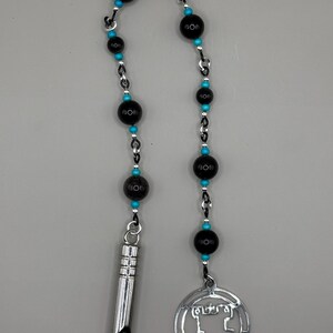 May include: A silver chain necklace with black beads and turquoise accents. The necklace has a silver whistle charm and a silver pendant with a geometric design.
