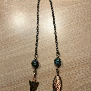 May include: A long, gold chain with two decorative beads and two charms. One charm is a small arrow and the other is an oval with an intricate design.