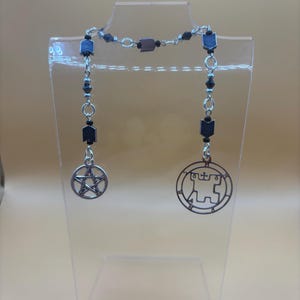 May include: A silver-toned necklace with a dark chain and dangling charms. The charms include a pentagram and a circular design. The necklace is displayed on a clear stand against a neutral background.