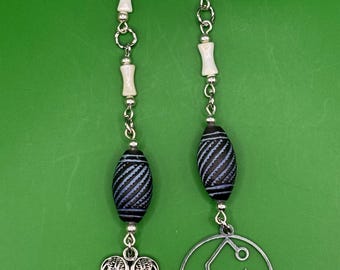 May include: A silver chain with white beads and black and blue striped beads. The chain has two charms attached to it, one is a pair of angel wings and the other is a circle with a symbol inside.