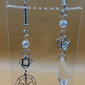 May include: Two decorative crystal suncatchers with silver-toned metal accents. One features a geometric design with a circular emblem, the other a faceted crystal pendant. Both have clear crystal beads and are suspended from a magnetic clasp.