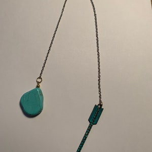 May include: A turquoise stone and arrow charm on a silver chain. The stone is a natural, irregular shape. The arrow is a stylized design with a pointed tip and a feathered tail.