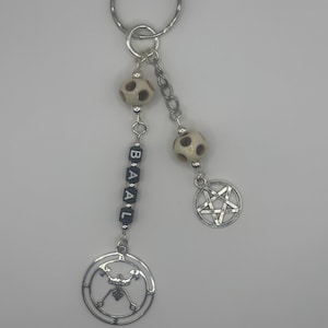 May include: A silver-toned keychain featuring a split design. One strand has black letter beads spelling BAAL and a circular sigil charm. The other strand has a silver pentagram charm. Both strands include cream-colored beads with dark spots.