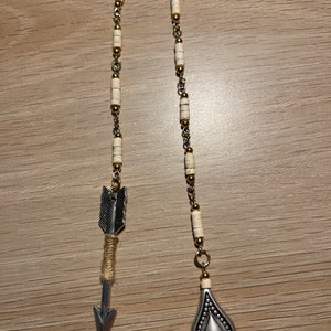 May include: A silver arrow charm with a brown twine wrap hangs from a chain with white and gold beads. A silver teardrop pendant with a textured design hangs from the chain.