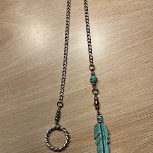May include: A silver-toned chain with a twisted ring and a turquoise feather charm. The chain is connected to a series of beads and a decorative feather. The jewelry is designed to be worn as a necklace or a keychain.