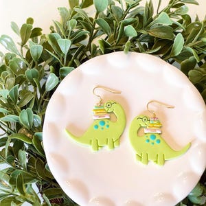May include: A pair of lime green dinosaur earrings with gold-colored hooks. Each earring features a dinosaur wearing glasses and carrying a stack of books. The dinosaurs have blue spots and are set on a white plate.