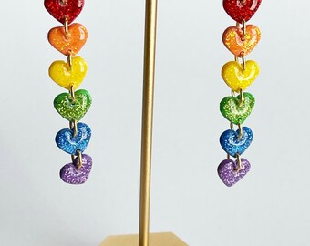 Könnte beinhalten: Ein Paar baumelnder Ohrringe mit herzförmigen Anhängern in Regenbogenfarben. Jedes Herz hat eine andere Farbe, darunter Rot, Orange, Gelb, Grün, Blau und Lila, mit Glitzerakzenten. Die Ohrringe hängen an goldfarbenen Haken.
