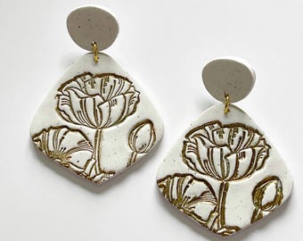 May include: A pair of white ceramic earrings with gold floral designs. The earrings are shaped like a diamond and have a textured surface.
