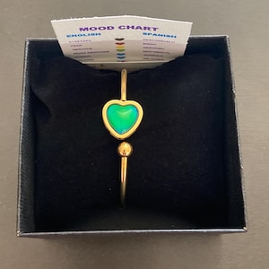 May include: A gold bracelet with a mood ring in the shape of a heart. The ring is green and iridescent. The bracelet is in a black box with a mood chart card that shows the different colors the ring can change to and what they mean.