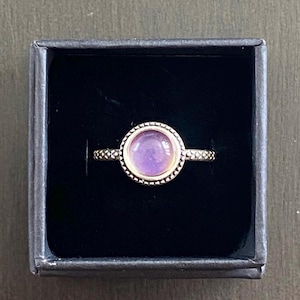May include: A silver ring with a round, iridescent purple stone set in a beaded bezel. The ring is displayed in a black jewelry box, showcasing its intricate design and color.