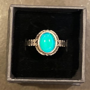 May include: A silver-toned ring with an oval, turquoise-colored stone. The ring is set in a decorative bezel with a detailed band, presented in a black box. The stone's color shifts from light to dark.
