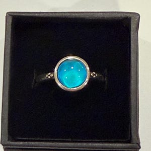 May include: A silver ring with a large, round, turquoise-colored stone. The stone is set in a silver bezel and the band has small decorative details. The ring is displayed in a black box.