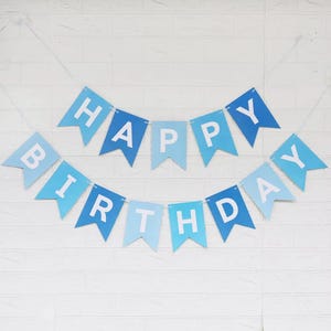 May include: A blue and light blue "Happy Birthday" banner with white lettering. The banner is made of pennant-shaped flags strung together with a white string. The banner is hanging against a white brick wall.