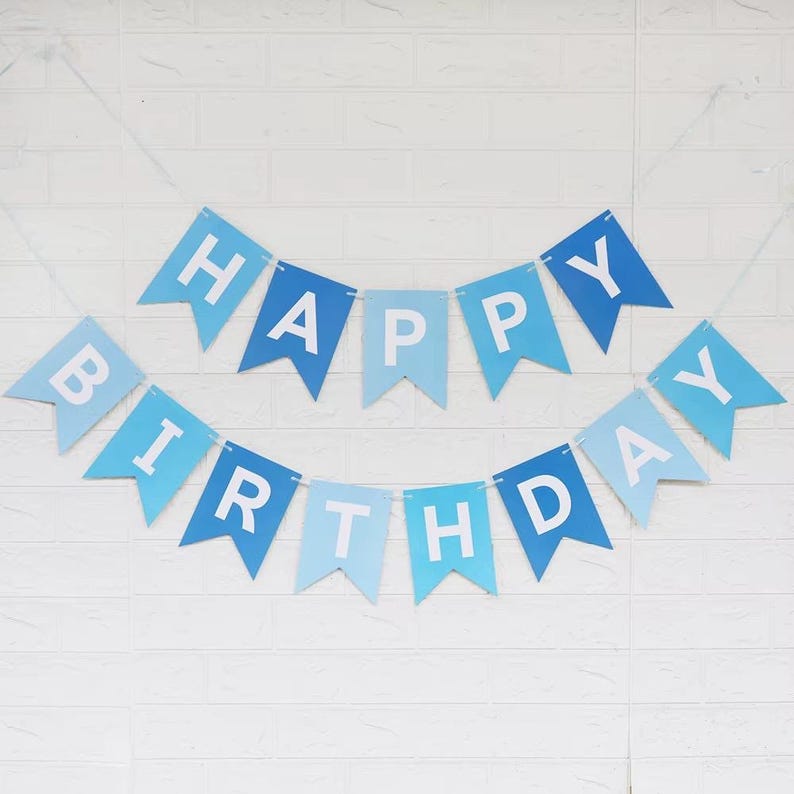 May include: A blue and light blue "Happy Birthday" banner with white lettering. The banner is made of pennant-shaped flags strung together with a white string. The banner is hanging against a white brick wall.